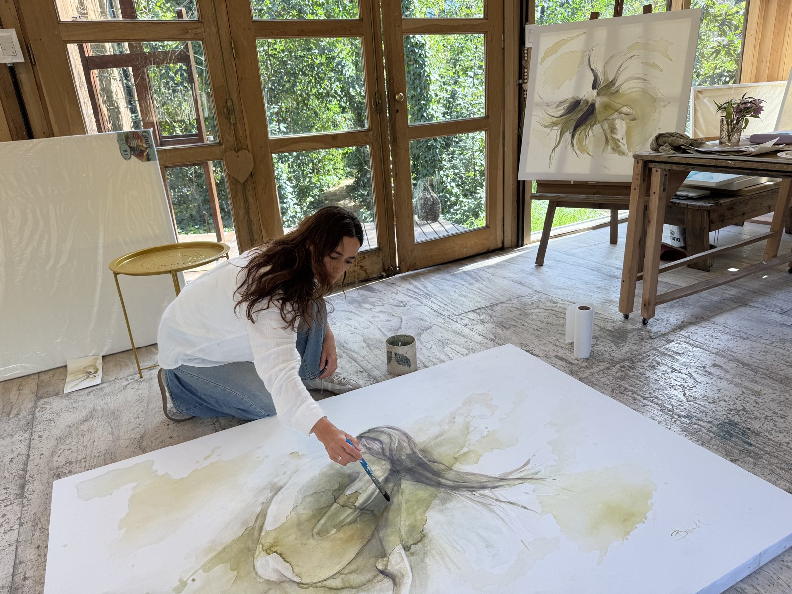 Brenda Rodríguez kneeling on her studio floor painting a large canvas, a finished work on the easel behind her
