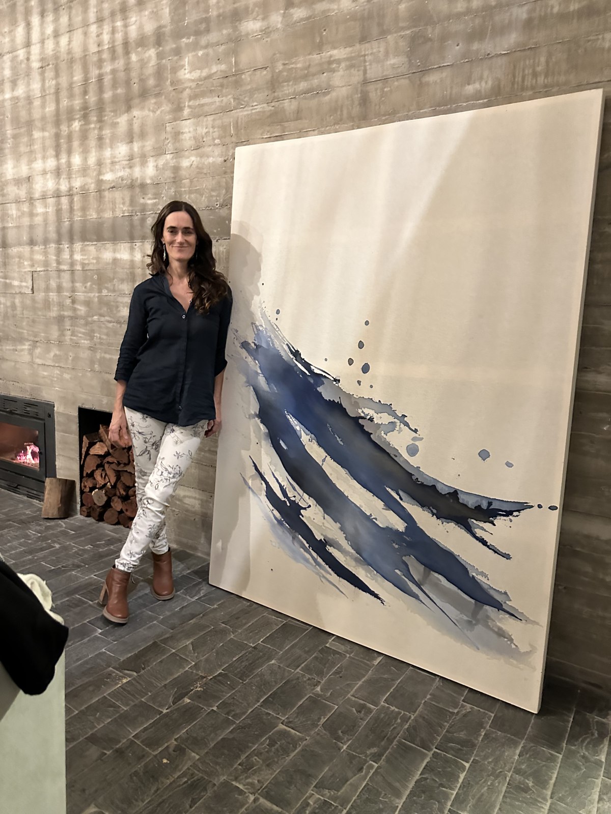 Brenda Rodríguez standing beside her large blue wave painting against a concrete wall